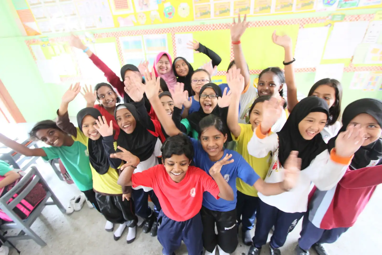 A class of pupils in coloured sports shirts waving and cheering with their teacher in a brightly decorated classroom