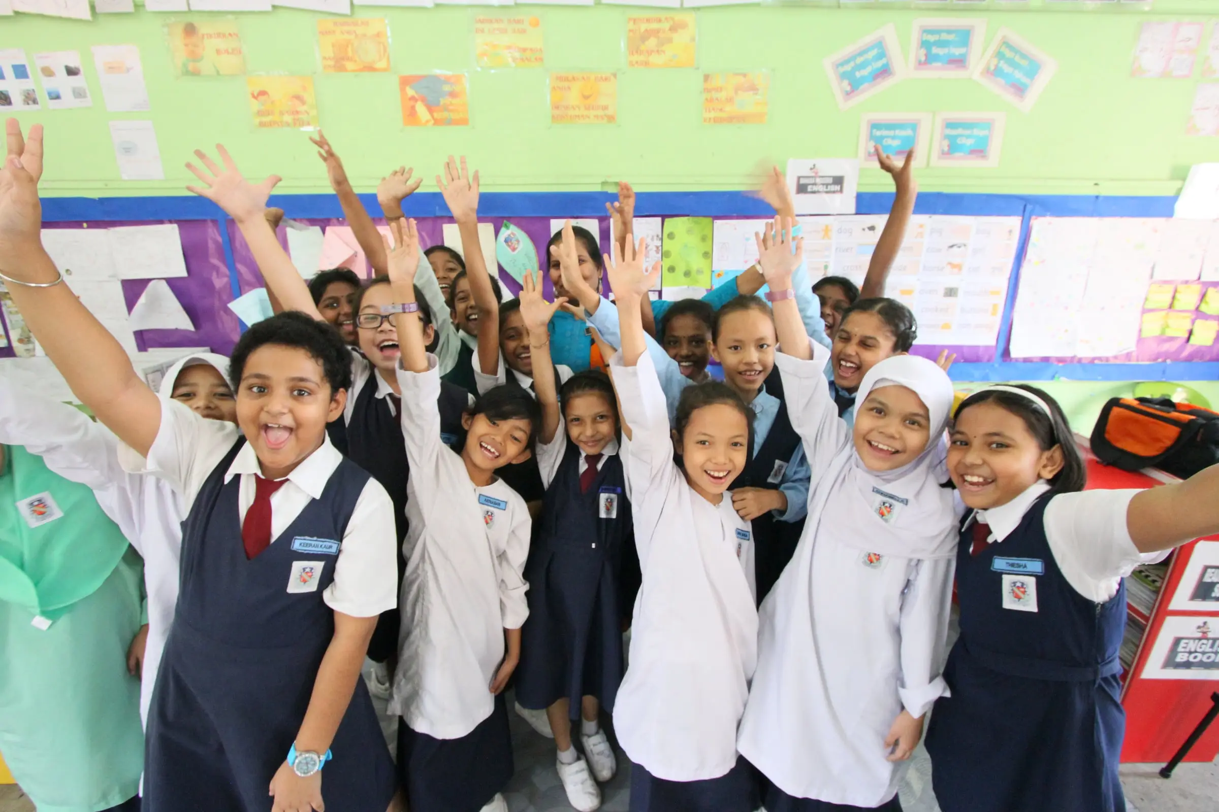 Primary pupils in school uniform raising their hands and smiling in class