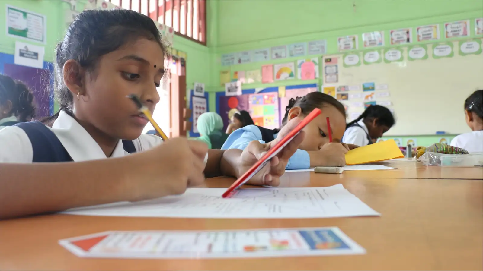 A primary pupil concentrating as she writes at her desk, her class working behind her