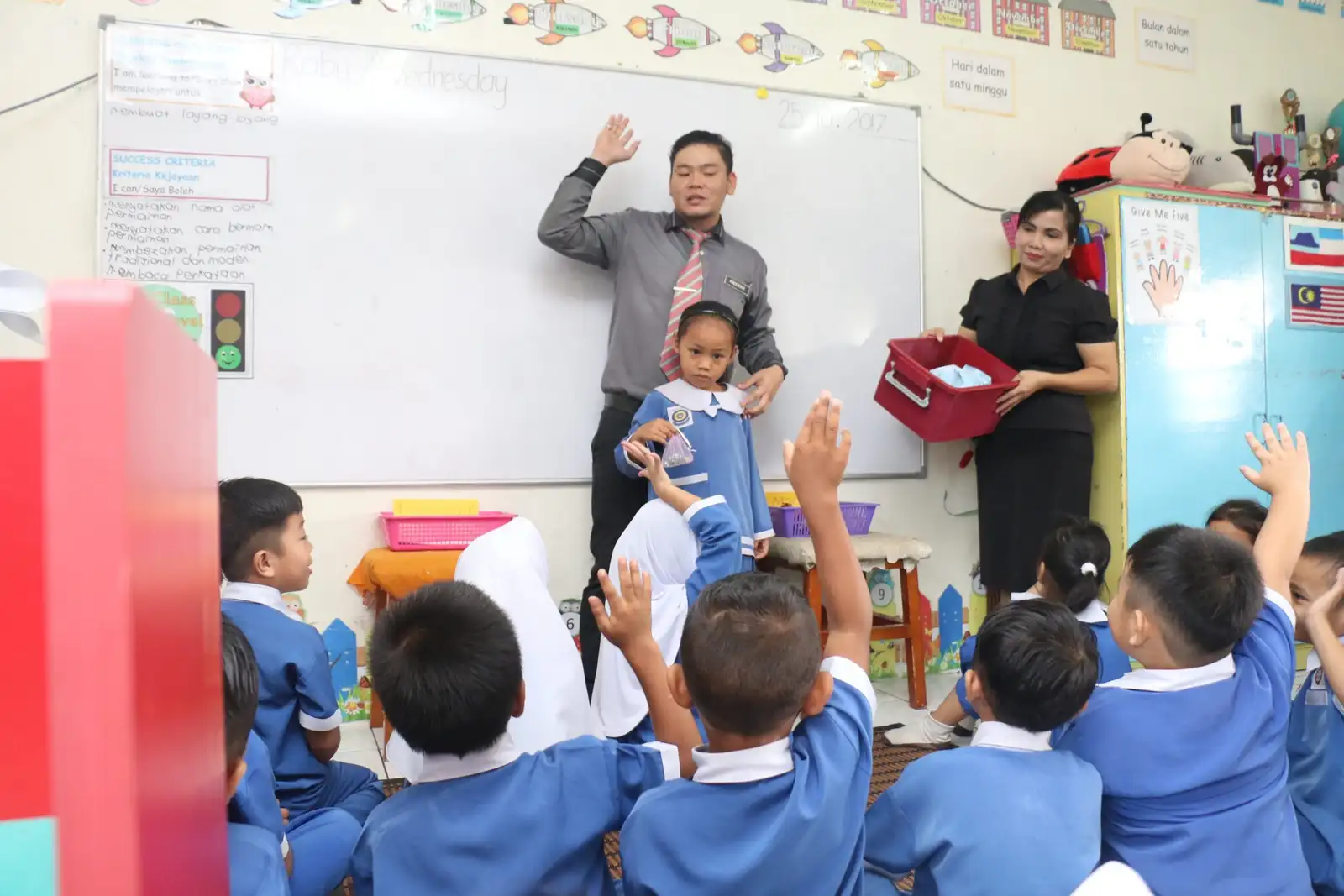 A teacher at the whiteboard with a pupil beside him as the class raise their hands