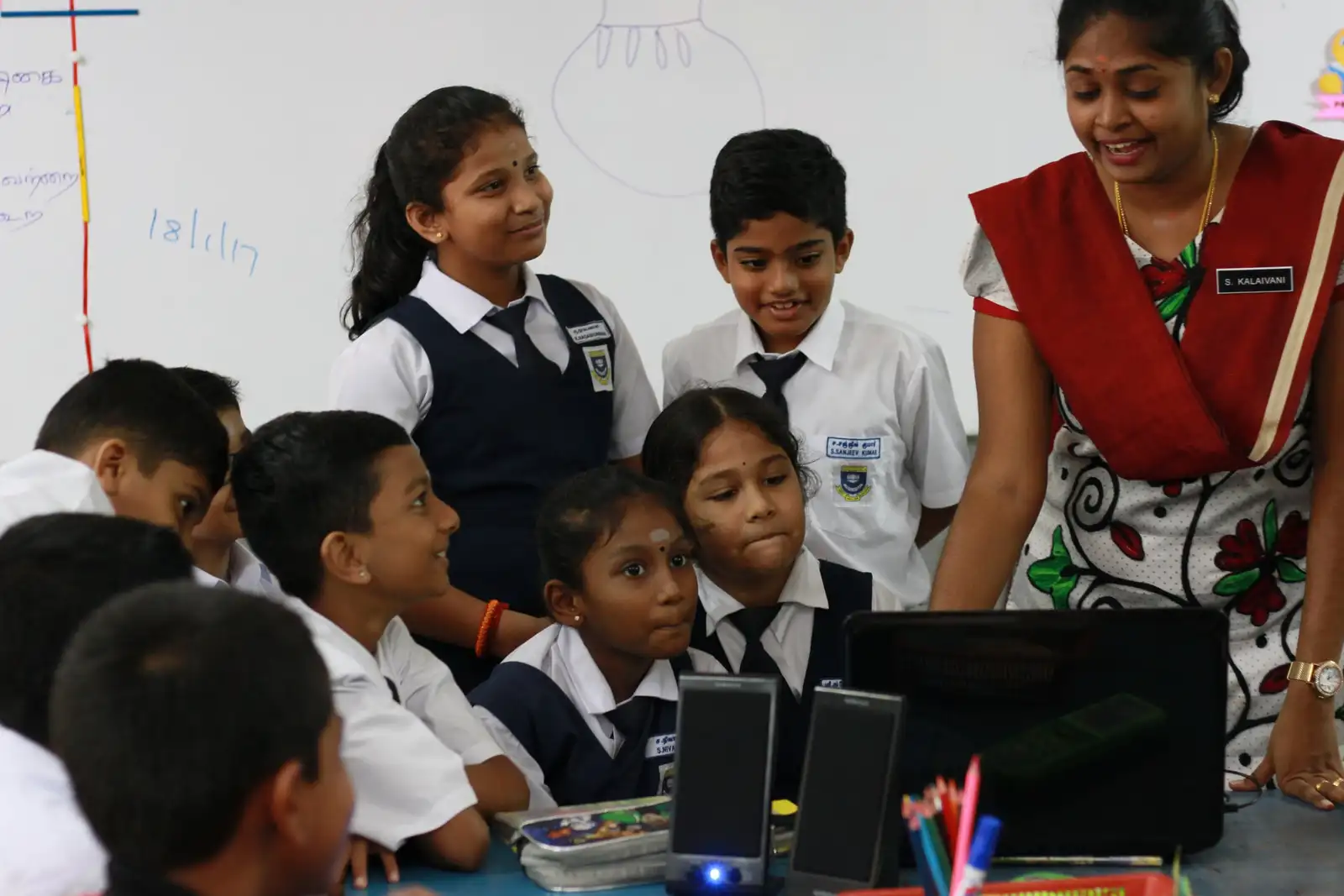 A teacher smiling down at a group of pupils gathered round a laptop in class