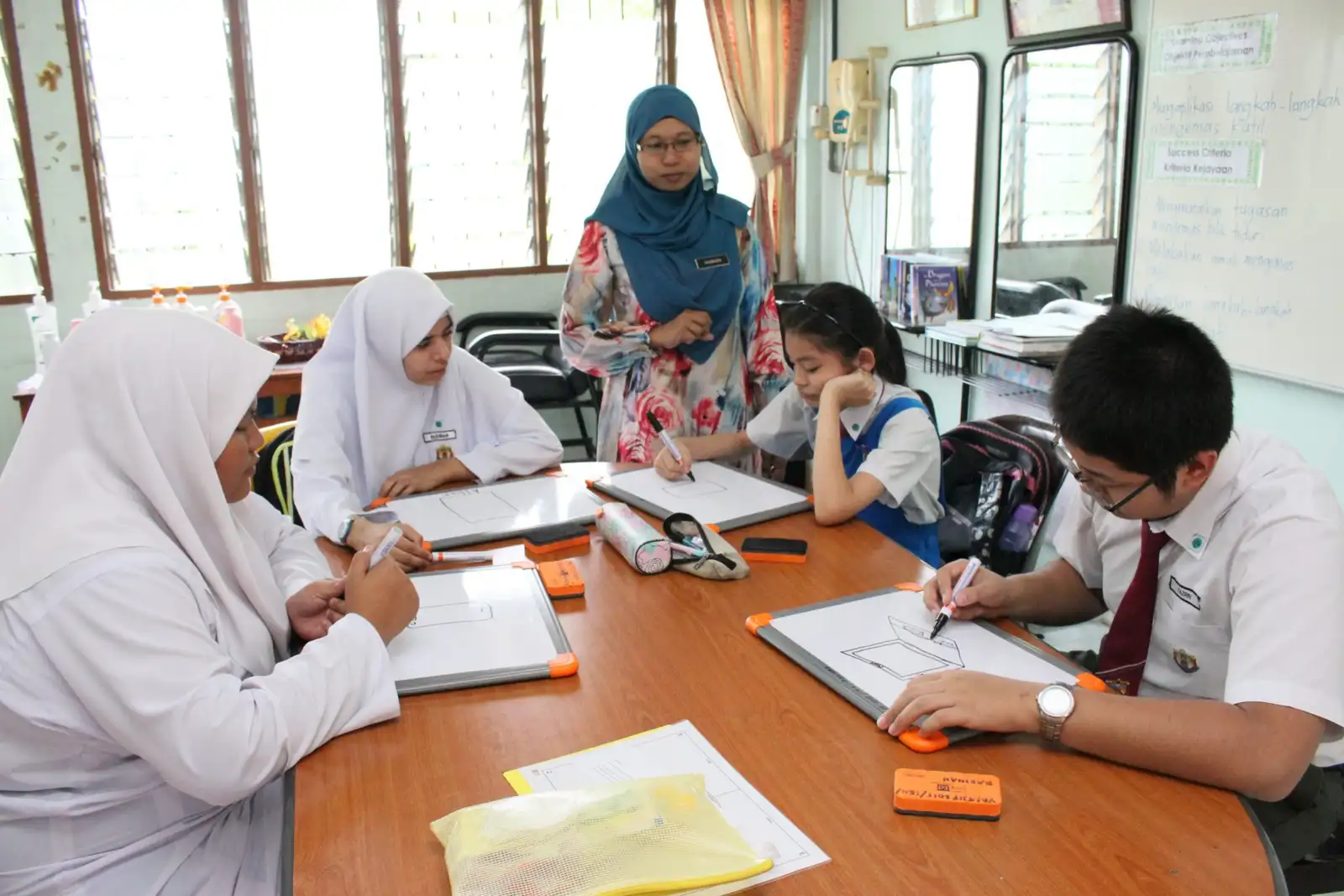 A teacher watching as pupils and teachers draw on small whiteboards around a classroom table